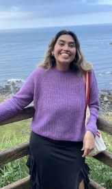 Picture of a woman smiling with the ocean and a green hillside in the background. She is leaning against a wooden fence and wearing a purple sweater and black skirt. She has brown hair with highlights that goes past her shoulders and caramel-colored skin. 